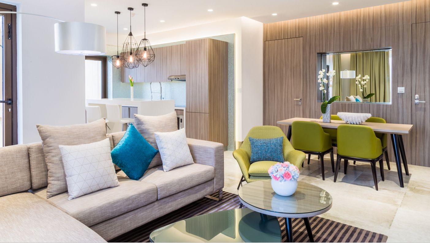 Modern open-plan living room with beige sofa, teal cushions, and dining area in soft tones.